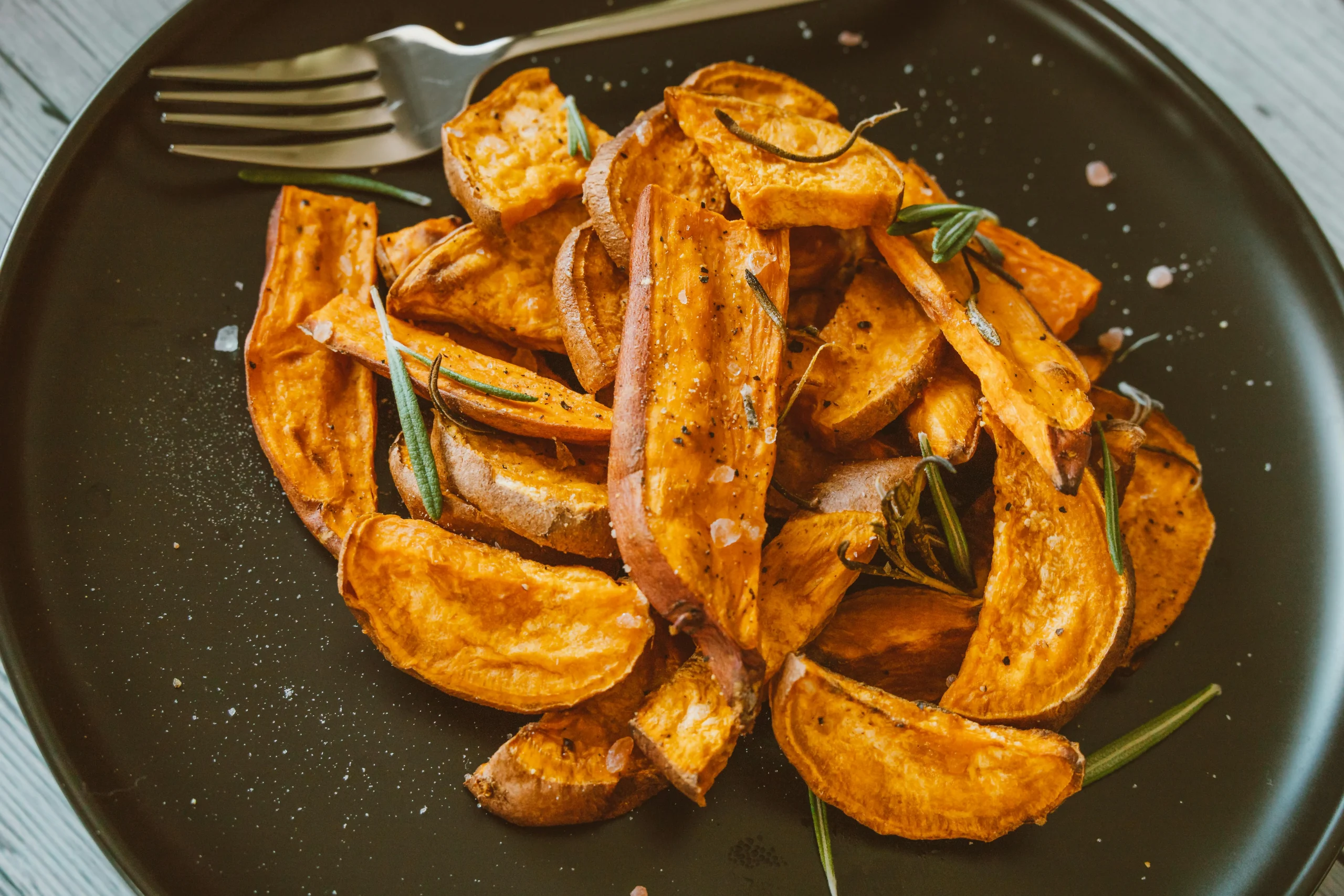 Roasted sweet potato wedges garnished with rosemary on a black plate.