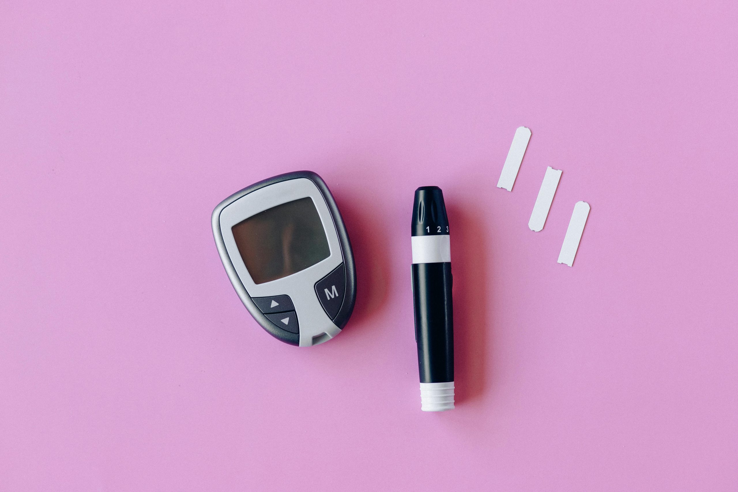 Diabetes tools like a glucose meter displayed on a pink background, illustrating how often to replace glucometer test strips.